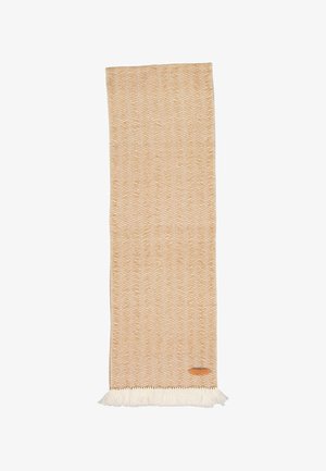 Tapis rectangulaire beige tissé avec un motif zigzag blanc, bords frangés et petite étiquette en cuir marron dans le coin inférieur droit.