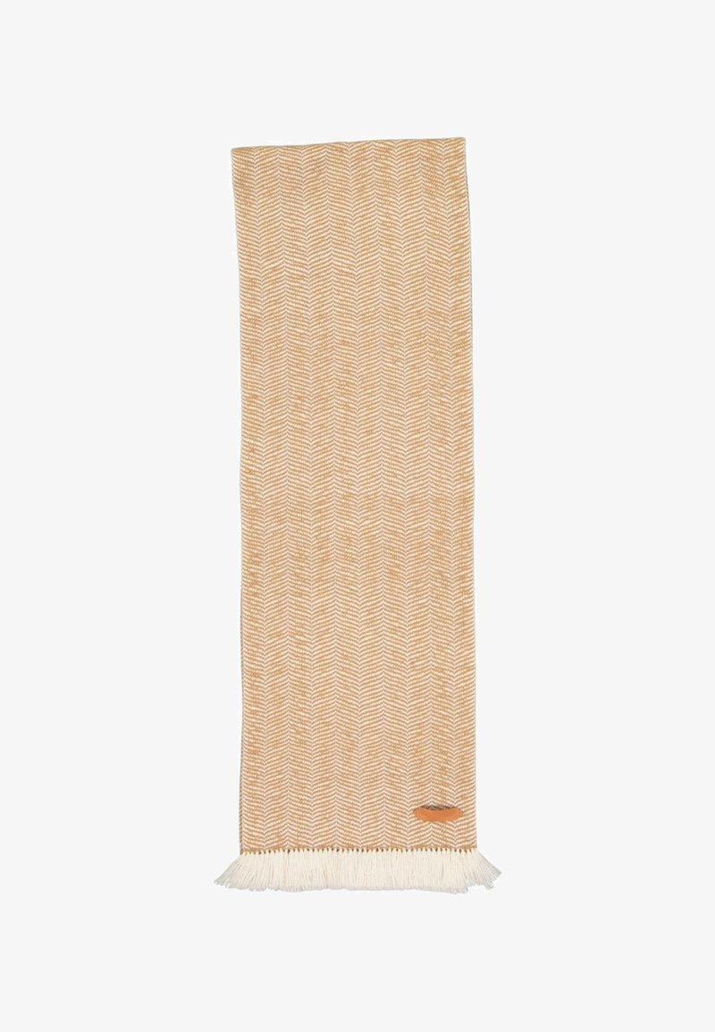 Tapis rectangulaire beige tissé avec un motif zigzag blanc, bords frangés et petite étiquette en cuir marron dans le coin inférieur droit.