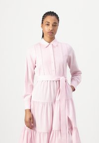 Vestido rosa com mangas longas, gola, detalhes plissados e cintura atada. A saia apresenta camadas franzidas e uma textura suave.