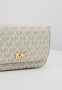 Sac à main beige et marron à motif avec un design répétitif du logo MK et un emblème MK doré sur le rabat avant sur un fond uni.