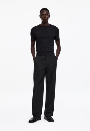 Homme grand debout les mains dans les poches, portant une chemise noire ajustée à manches courtes, un pantalon noir large et des chaussures habillées noires sur fond blanc.