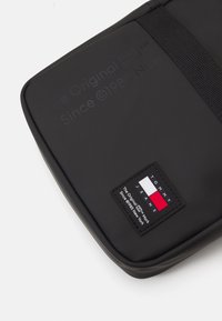 Czarna torba na laptopa o gładkiej fakturze, z naszywką z logo w czerwonym, białym i niebieskim kolorze, a także wytłoczonym tekstem marki na powierzchni.