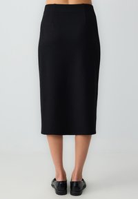 Vrouw in een aansluitende zwarte rok tot op de knie en zwarte leren instapschoenen, staande tegen een effen lichtgrijze achtergrond.