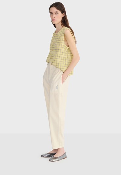 Femme debout de profil portant un haut sans manches à carreaux jaunes, un pantalon crème taille haute avec détail de poche, et des ballerines argentées.