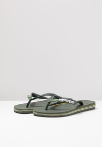 Un paio di flip-flop Havaianas verde oliva con dettaglio della bandiera brasiliana e logo bianco sulle cinghie, strisce blu e gialle sui suole.