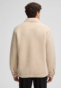 Beige fleece jacket with a textured surface, oversized fit, and a turn-down collar. Features button accents at the cuffs.