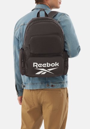 Mann trägt eine Jeansjacke und beige Hose, trägt einen schwarzen Reebok-Rucksack mit mehreren Reißverschlussfächern.