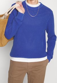 Pull en maille côtelée bleue associé à une chemise blanche et un pantalon beige. Un sac fourre-tout texturé de couleur moutarde pend du bras.