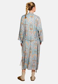 Lange, blauw gestreepte robe met paisley ontwerp, zachte stof, tailleband en brede mouwen; gedragen met comfortabele beige sokken en slippers.