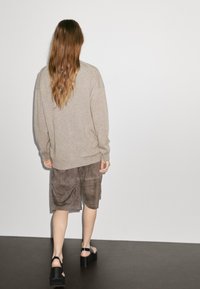 Oversized beige tröja, bruna mockashorts och svarta platåsandaler. Rakt hår faller över ryggen i lösa vågor. Minimalistisk bakgrund.