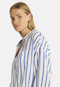 Giovane donna con lunghi capelli biondi, che indossa una camicia a righe blu e bianche, sorride leggermente con un trucco delicato e orecchini a cerchio d'oro.