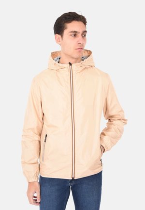 Uomo che indossa una giacca beige con cappuccio e cerniera frontale, con le mani nelle tasche, guarda verso sinistra.