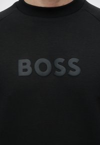 Άτομο που φοράει μαύρο φούτερ με τη λέξη BOSS τυπωμένη με μεγάλα ανάγλυφα μαύρα γράμματα στο στήθος.