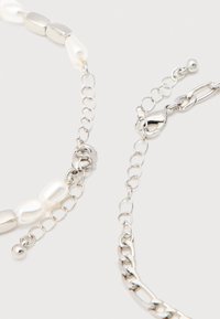 Δύο ασημένια βραχιόλια αλυσίδας με κούμπωμα αστακού και ένα με λευκά μαργαριτάρια και ασημένιες χάντρες σε ανοιχτό φόντο.