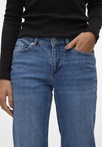 Jeans denim biru dengan potongan klasik, dilengkapi lima kantong, penutupan kancing, dan jahitan yang terlihat. Dipakai dengan atasan lengan panjang berwarna hitam.