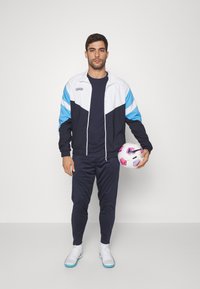 Marine trainingspak met witte en lichtblauwe accenten, bestaande uit een jack met rits en slim-fit broek. Houdt een meerkleurige voetbal vast.