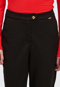 Zwarte broek met een gladde textuur, een getailleerd design en een gouden knoopaccent in de taille. Het model draagt een rode top met lange mouwen.