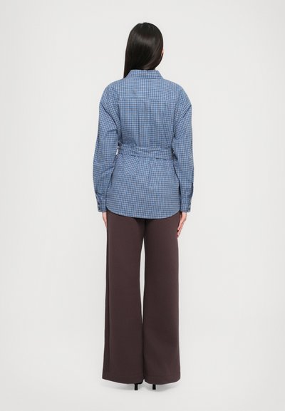 Sinine ruuduline särk, millel on pehmendatud vöökoht ja pikad varrukad, koos laia sigarettpüksiga pruunides toonides, vaadates tagant.