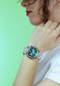 Reloj de pulsera plateado con una esfera verde, acentuado por un bisel engastado con cristales y una pulsera de acero inoxidable. El fondo es de color verde claro.