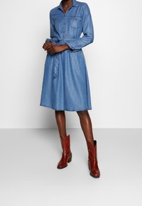 Jeansklänning med bälte i midjan, långa ärmar och två bröstfickor. Bärs med röda lädercowboystövlar och en midellång nederkant.