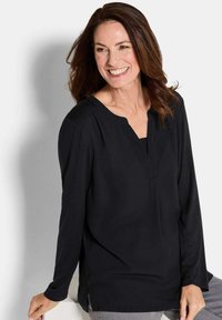 Donna sorridente con capelli castani indossa una blusa nera a maniche lunghe e pantaloni grigio chiaro, seduta e appoggiata su una superficie bianca contro uno sfondo bianco.