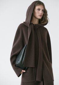 Jeune femme portant un manteau en laine marron à capuche, tenant un sac à main en cuir noir sur un fond uni.