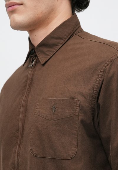 Camisa castanha de botão com colarinho pontiagudo, tecido texturizado e um bolso no peito com um logótipo bordado. O ajuste parece ser ajustado.