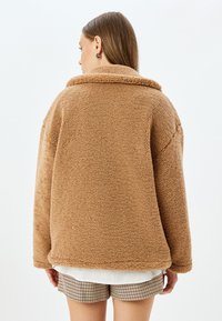 Giacca in pile marrone, morbida e voluminosa, con colletto e vestibilità oversize. Tessuto soffice con una leggera lucentezza, presenta cuciture a vista e orlo arrotondato.