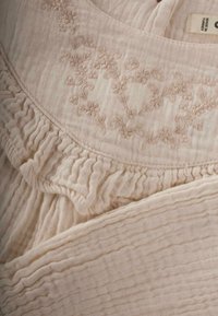 Helle beige Textilie mit zarter floraler Stickerei und Rüschensaum. Der Stoff hat eine strukturierte, zerknitterte Oberfläche.