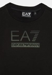 Juodas medvilninis marškinėliai su blizgiu EA7 logotipu ir kontrastinga pilka "EMPORIO ARMANI" užrašu. Klasikinis apvalus iškirpimas, lygus paviršius.