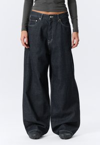 Mørkeblå, vid jeans med høy midje, med lyse sømmer, frontlommer og knappelukking. Tykt, strukturert stoff.