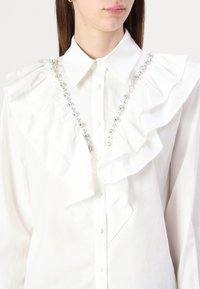 Blusa de algodón blanca con volantes prominentes en la parte delantera y una fila de gemas transparentes decorativas en el cuello. Cierre clásico de botones.