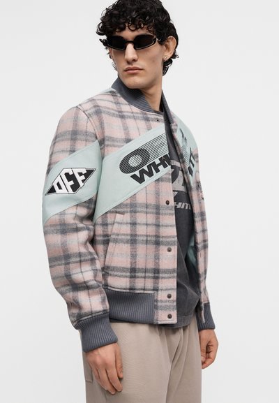 Jeune homme portant des lunettes de soleil noires et une veste à carreaux rose-gris avec des patchs bleu clair et un texte graphique, debout devant un fond uni.