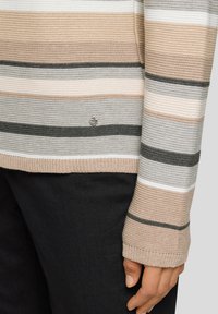 Beigefarbene, graue, weiße und schwarze gestreifte Strickpulloverärmel und -torso mit einem kleinen herzförmigen "mit Liebe gemacht"-Etikett, getragen mit schwarzen Hosen.