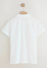 Weißes Poloshirt, das an einem Holzknopf an einer hellen Wand hängt, mit einem schlichten Kragen und kurzen Ärmeln.