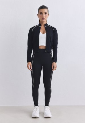 Mujer de pie contra una pared lisa, vestida con una chaqueta negra con cremallera, leggings negros con rayas blancas, top corto blanco y zapatillas blancas.