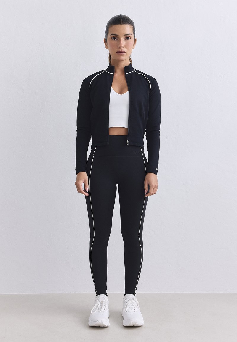 Femme debout contre un mur uni, portant une veste noire zippée, un legging noir avec des rayures blanches, un crop top blanc et des baskets blanches.