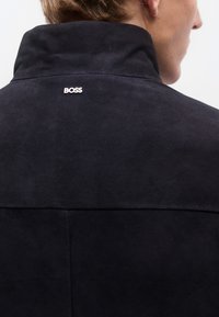 Tengerészkék színű nadrágszárbőr dzseki magas gallérral, hátul ezüst "BOSS" logóval. Sima textúrájú és minimalista kialakítású.