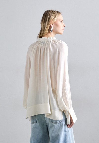 Blusa crema ampia con colletto arricciato, maniche lunghe e orlo leggermente asimmetrico, abbinata a jeans in denim azzurro chiaro.