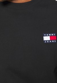 Czarna bluza z bawełny z prążkowanym dekoltem. Na piersi znajduje się naszywka z logo Tommy Jeans z niebieskimi, czerwonymi i białymi akcentami.