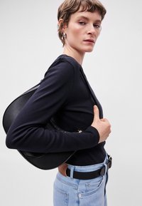 Μαύρη δερμάτινη τσάντα ώμου με στρογγυλό σχήμα, υφή, χρυσές λεπτομέρειες, φορεμένη πάνω από μπλε μακρυμάνικο τοπ, σε συνδυασμό με ανοιχτόχρωμα μπλε τζιν.