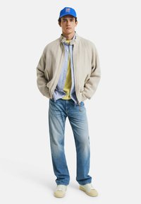 Lightweight linen bomber jacket in beige, layered over a green shirt and blue button-up. Paired with faded blue jeans and white sneakers. Blue cap.