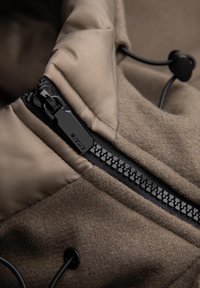 Detailní záběr na černý zip s poutkem na hnědém látkovém kabátu s polstrovaným límcem a nastavitelnou šňůrkou se stahovacími prvky.