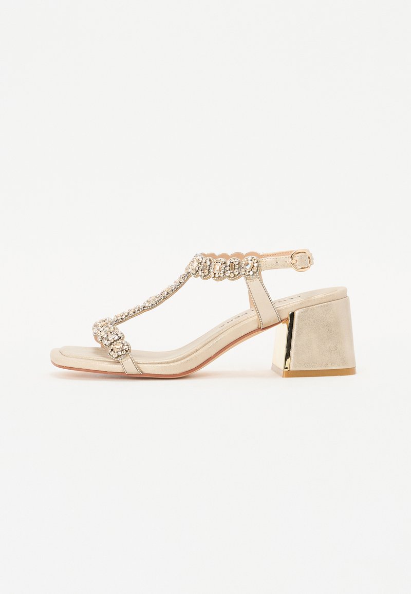 Goldene Sandale mit Blockabsatz, T-Riemen-Design und verzierten Strass-Akzenten an den Riemen. Glatte Textur und minimalistisches Profil.