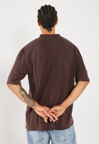 Bruine T-shirt met korte mouwen, met een relaxte pasvorm, ronde hals en een gestructureerde stof, gedragen met blauwe denim jeans. De achterzijde toont gevlochten haar.