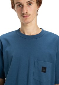 Blue cotton t-shirt with short sleeves, featuring a left chest pocket and a black logo patch. Smooth texture and relaxed fit.