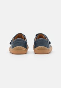 Zapatos de cuero navy con una textura suave, presentando una suela de goma color tostado y una correa de gancho y bucle. La vista trasera muestra un tacón redondeado y detalles de costura.