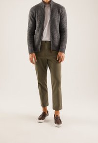 Man i grå zip-up stickad cardigan, vit t-shirt, olivgröna cropped byxor och bruna slip-on skor som står på vit bakgrund.