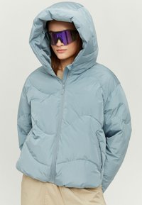 Light blue puffer jacket with a large hood, quilted design, front zipper, and side pockets. Model wearing reflective sunglasses.
