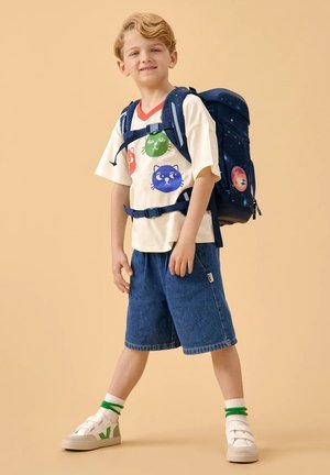 Young boy wearing a cat-print T-shirt, denim shorts, white sneakers, and a navy blue backpack with space-themed details.